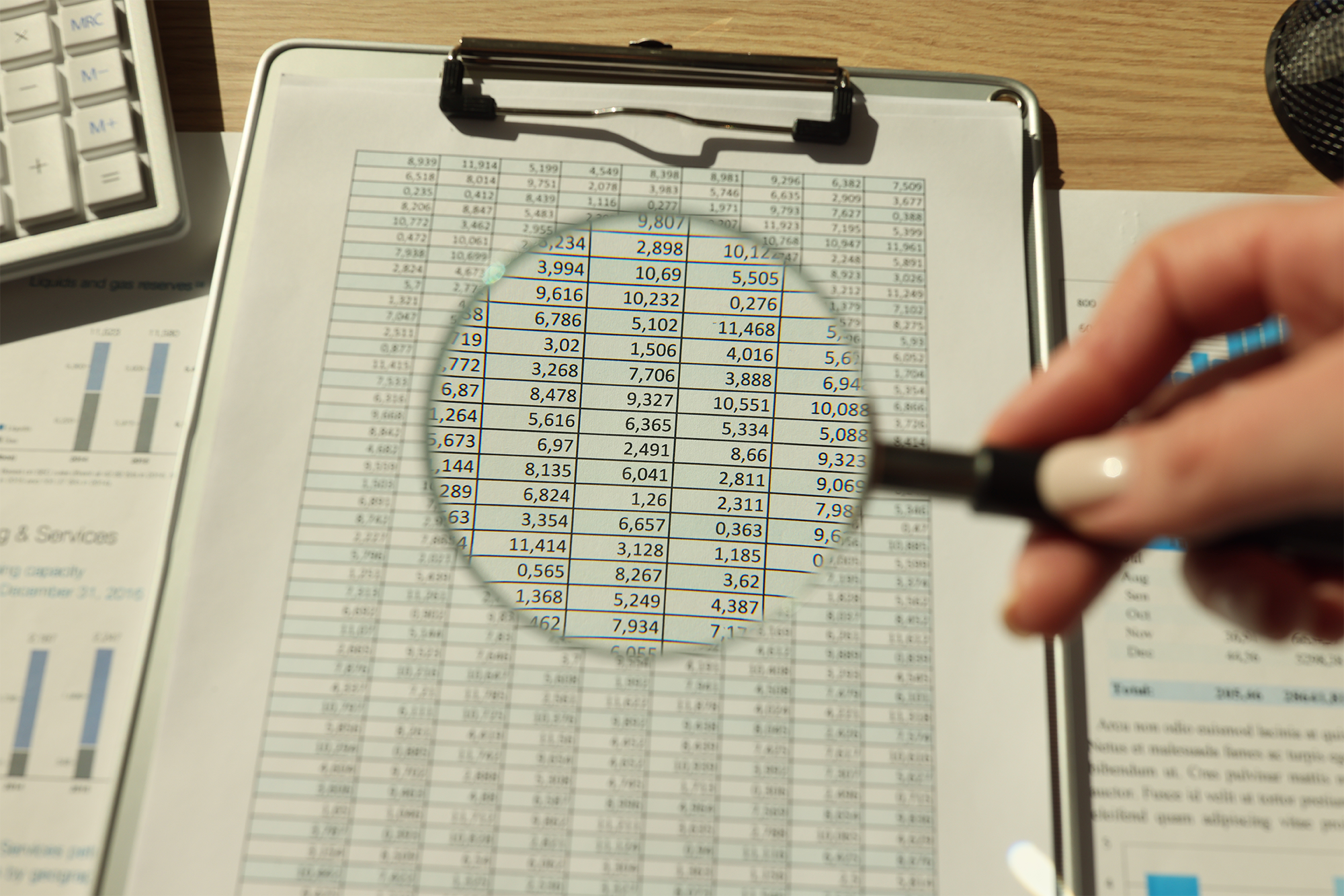 Mão de uma mulher segurando o vidro da lupa acima dos papéis contábeis na investigação da mesa