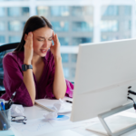 Jovem empresária sobrecarregada de trabalho que sofre de dor de cabeça no escritório sentada na mesa em frente ao monitor do computador com problemas com o trabalho