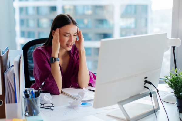 Jovem empresária sobrecarregada de trabalho que sofre de dor de cabeça no escritório sentada na mesa em frente ao monitor do computador com problemas com o trabalho